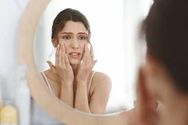 Inside-Out Care: Why True Beauty Sleep Starts with Stress Relief 7 A stressed woman looking at her skin reflection in a round bathroom mirror with a worried expression